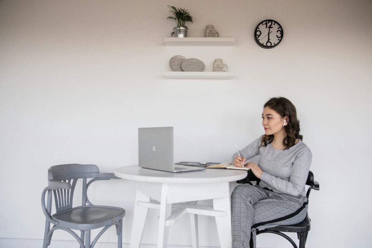 ethnic female student in earbuds writing on copybook near laptop