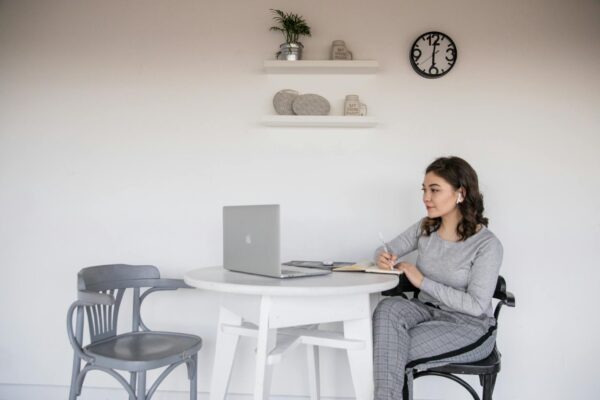 ethnic female student in earbuds writing on copybook near laptop